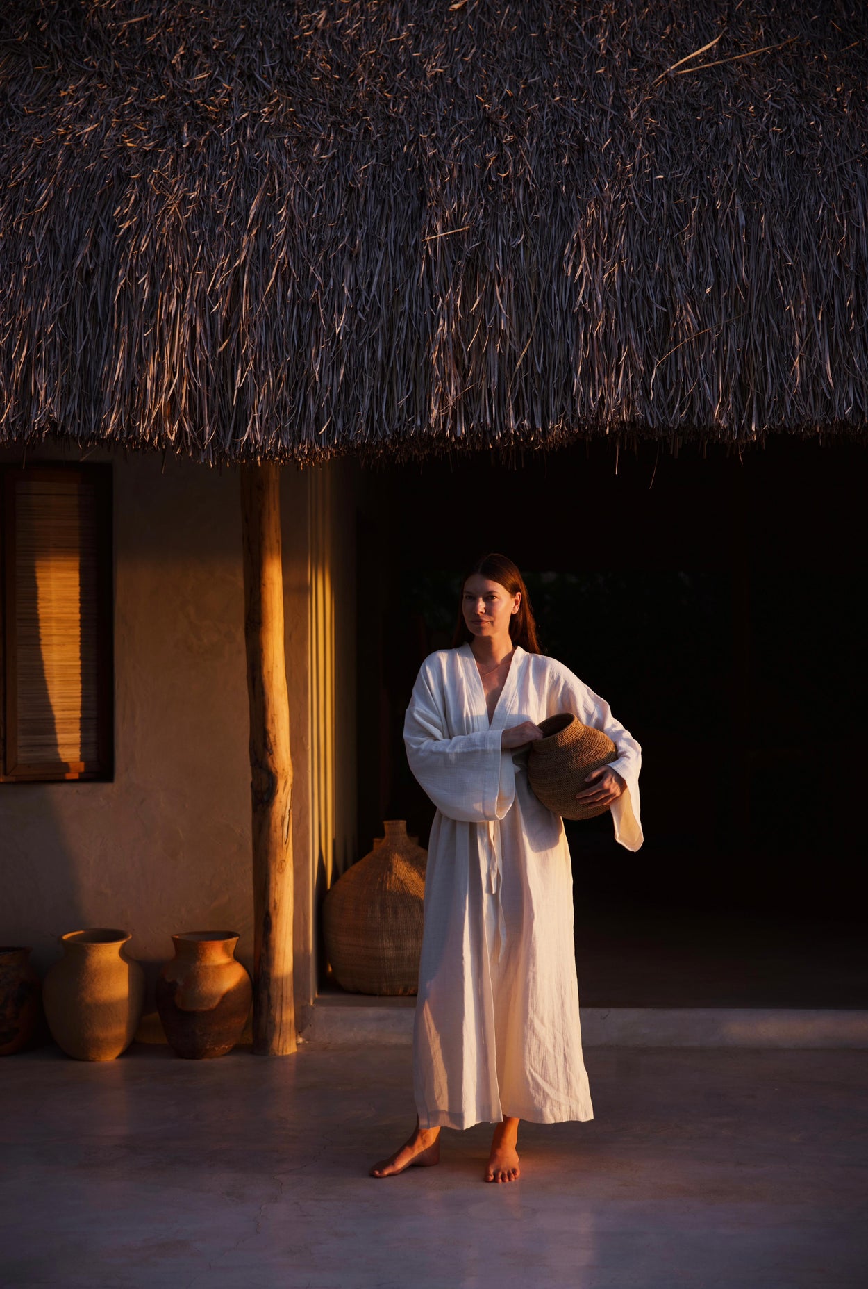 Oatmeal muslin robe by atelier an.nur photographed in warm golden-hour light as a woman holds a woven basket in front of an African hut.